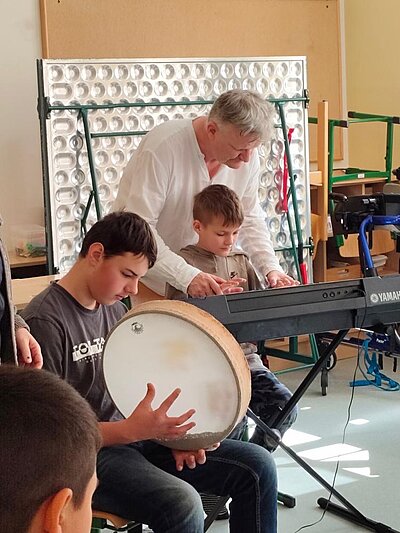 Im Vordergrund ein Schüler im Rollstuhl mit einem Rhythmusinstrument, der Musikpädagoge mit einem weitern Schüler am Keyboard im Hintergrund