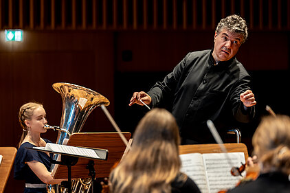 Dirigent mit erhobenen Armen vor einem Orchester mit Musiker*innen und Notenständern in einem Konzertsaal