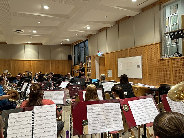 Dirigentin leitet die Probe eines großen Blasorchesters in einem Proberaum; die Musikerinnen und Musiker sitzen mit Instrumenten und Notenpulten im Halbkreis.