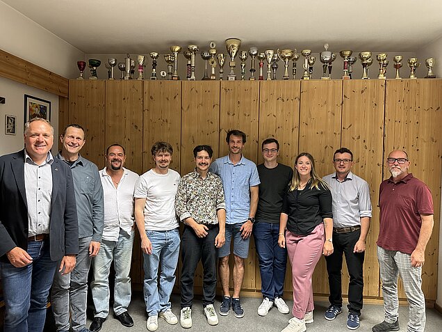 Gruppenfoto von zehn Personen aus dem Bereich Musikausbildung vor einem Holzschrank mit zahlreichen Pokalen.