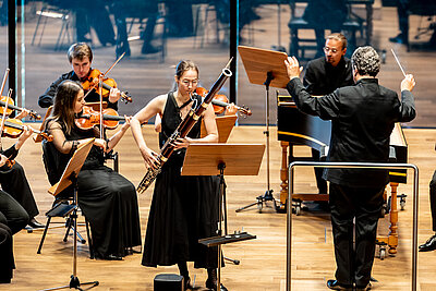 ein Teil des Orchesters, Dirigent und Wettbewerbskandidatin spielen live auf einer Konzertbühne