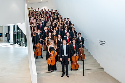 Das gesamte Orchester steht aufgereiht auf einer Treppe, im Vordergrund der Dirigent